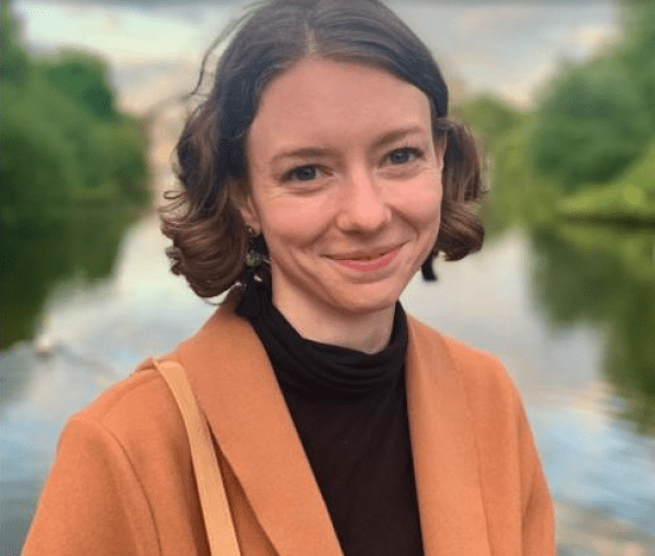 Portrait photo of Emma Satterfield in front of body of water. She is wearing a light orange blazer and smiling.