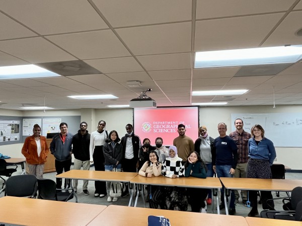 High school students and teachers pose with Associate Chair George Hurtt in front of a red GEOG backdrop in a LeFrak classroom