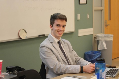 Picture of Blake Munshell. Blake is a white man with with blonde/brown hair, wearing a grey suit jacket and purple tie. He is sitting behind a desk with a laptop in front of him. 